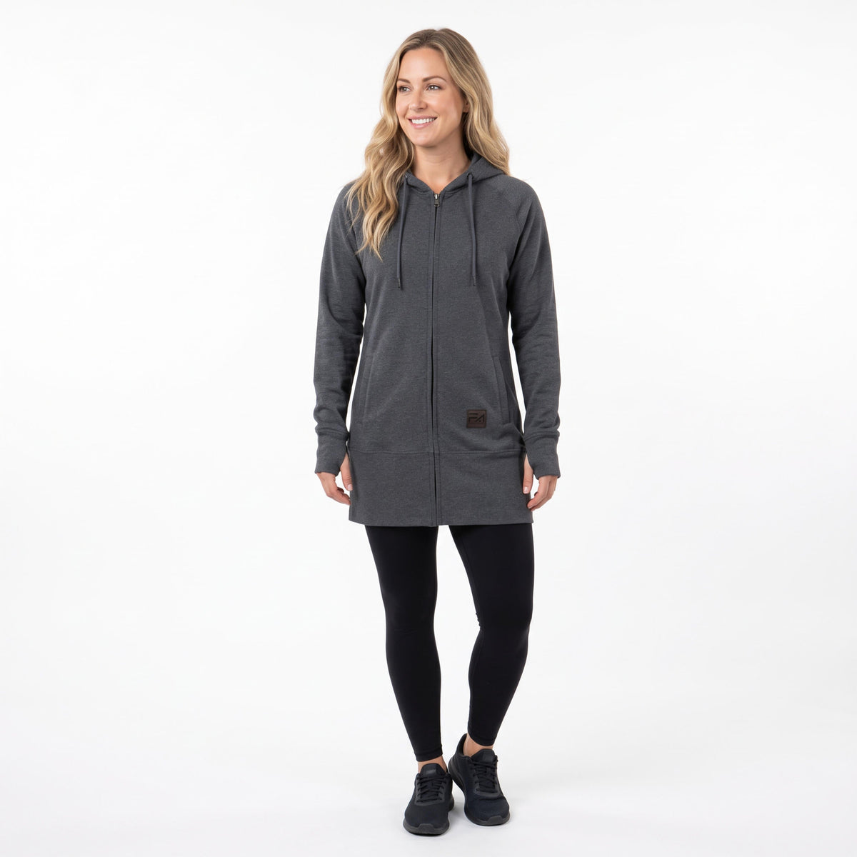 A woman modeling a dark gray zip-up hooded sweatshirt with black leggings and black sneakers, standing against a plain white studio background.