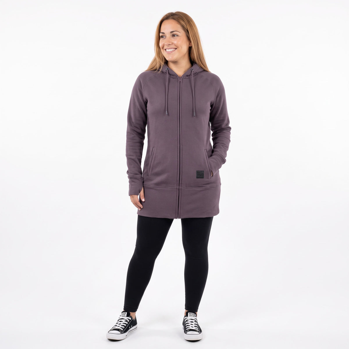 A woman modeling a dusty purple zip-up hoodie with black leggings and black sneakers, standing against a plain white studio background.