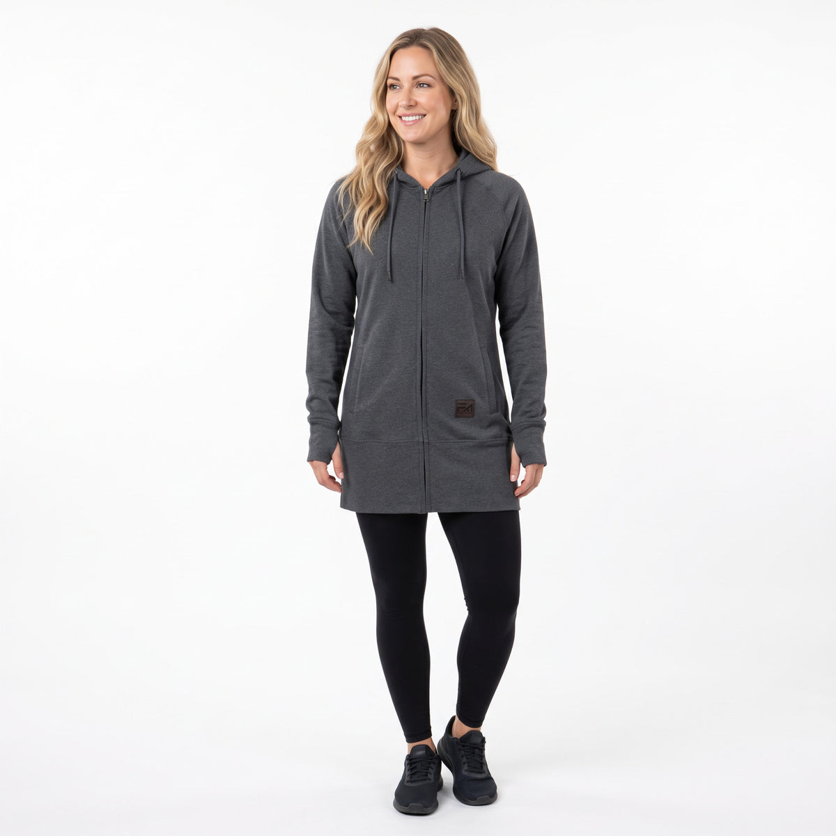 A woman modeling a dark gray zip-up hooded sweatshirt with black leggings and black sneakers, standing against a plain white studio background.