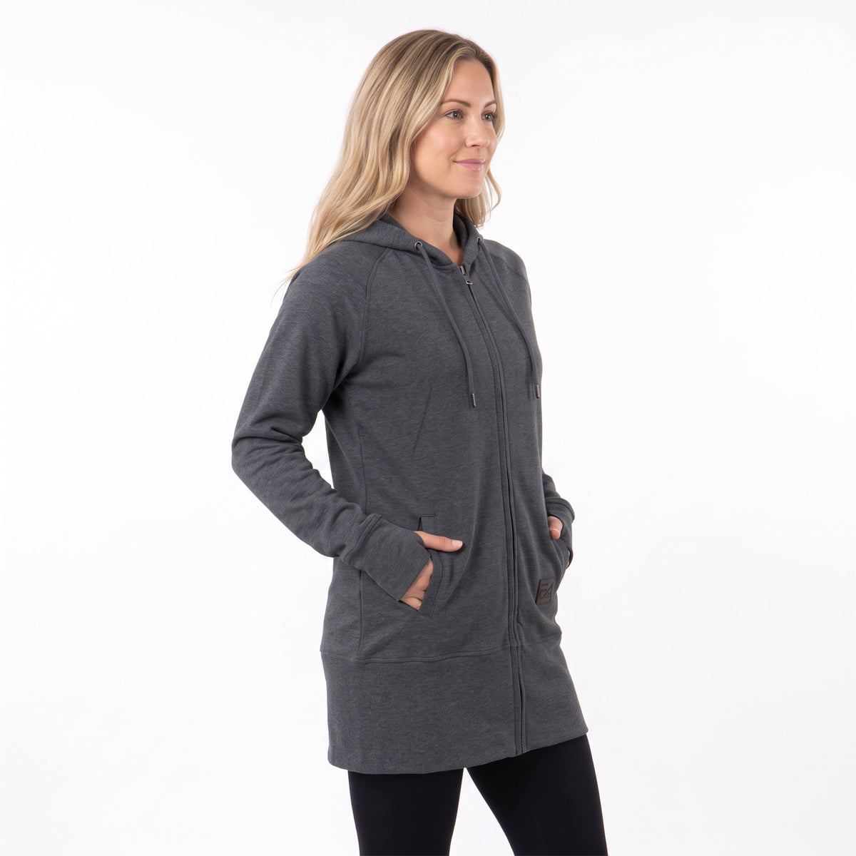 A woman modeling a dark gray zip-up hoodie with a drawstring hood and front pockets, paired with black leggings, against a plain white studio background.