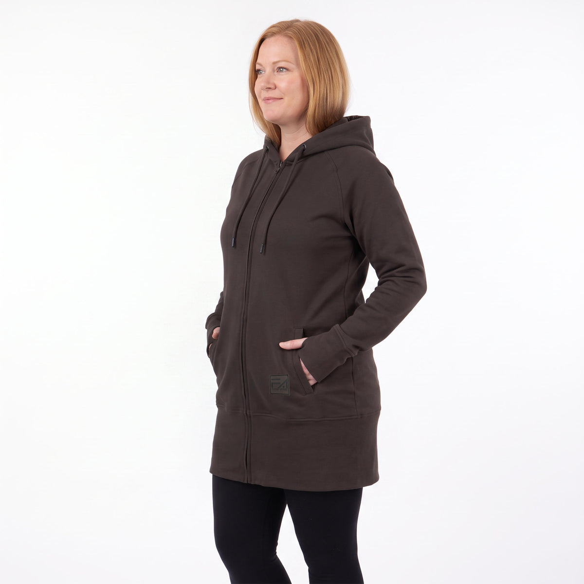 A woman wearing a dark brown zip-up hooded sweatshirt with side pockets, paired with black leggings, standing against a plain white studio background.