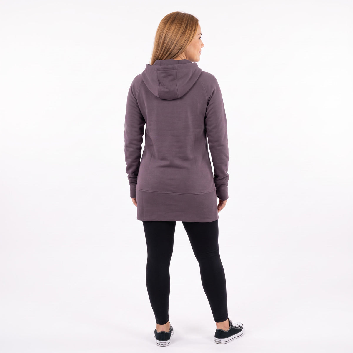 A woman viewed from the back wearing a dusty purple longline hoodie, black leggings, and black sneakers, standing against a plain white studio background.