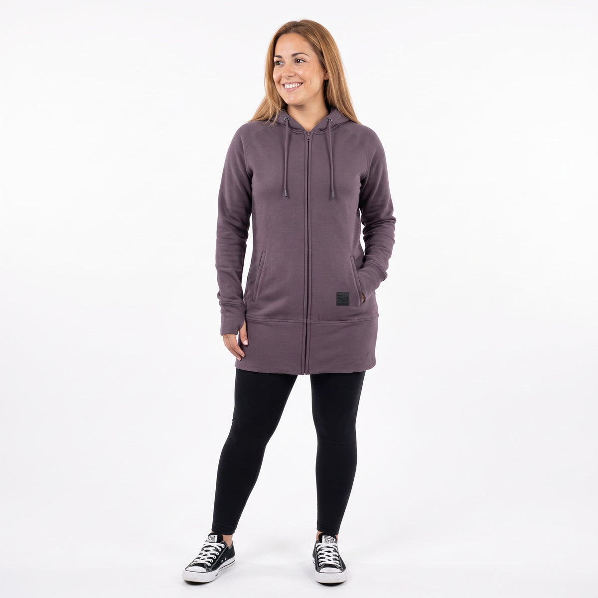 A woman modeling a dusty purple zip-up hoodie with black leggings and black sneakers, standing against a plain white studio background.