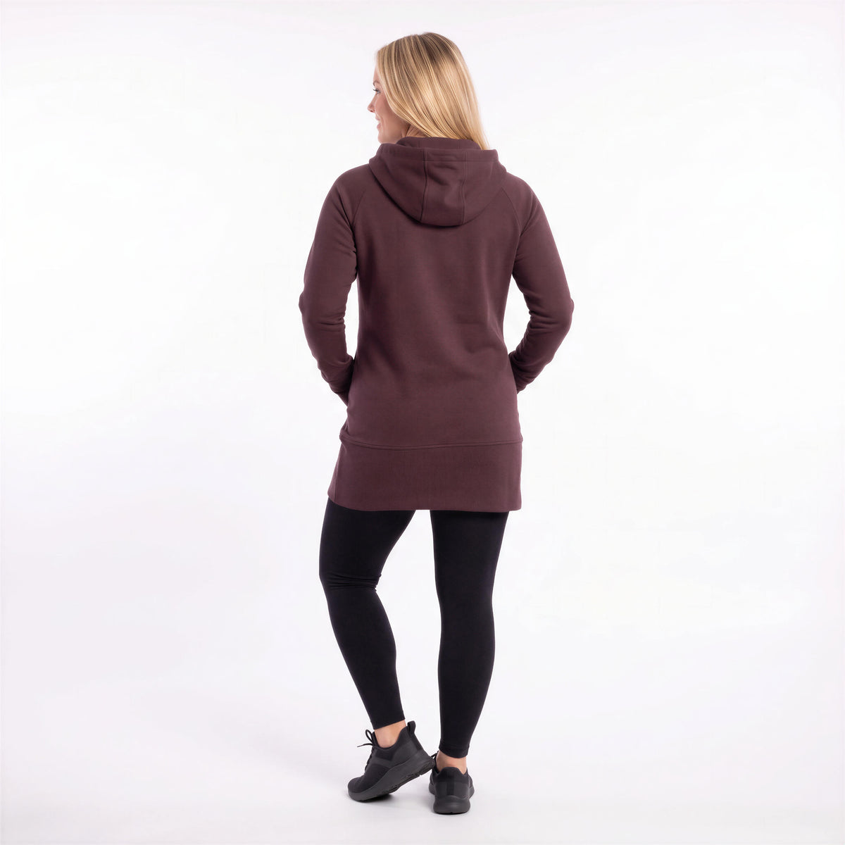 A woman is shown from behind wearing a dark plum-brown hooded sweatshirt, black leggings, and black sneakers against a plain white studio background.