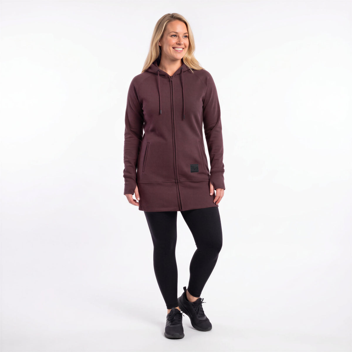 A woman modeling a dark plum-colored zip-up hoodie with black drawstrings and a small black brand patch on the lower front, paired with black leggings and black sneakers, against a plain white studio background.