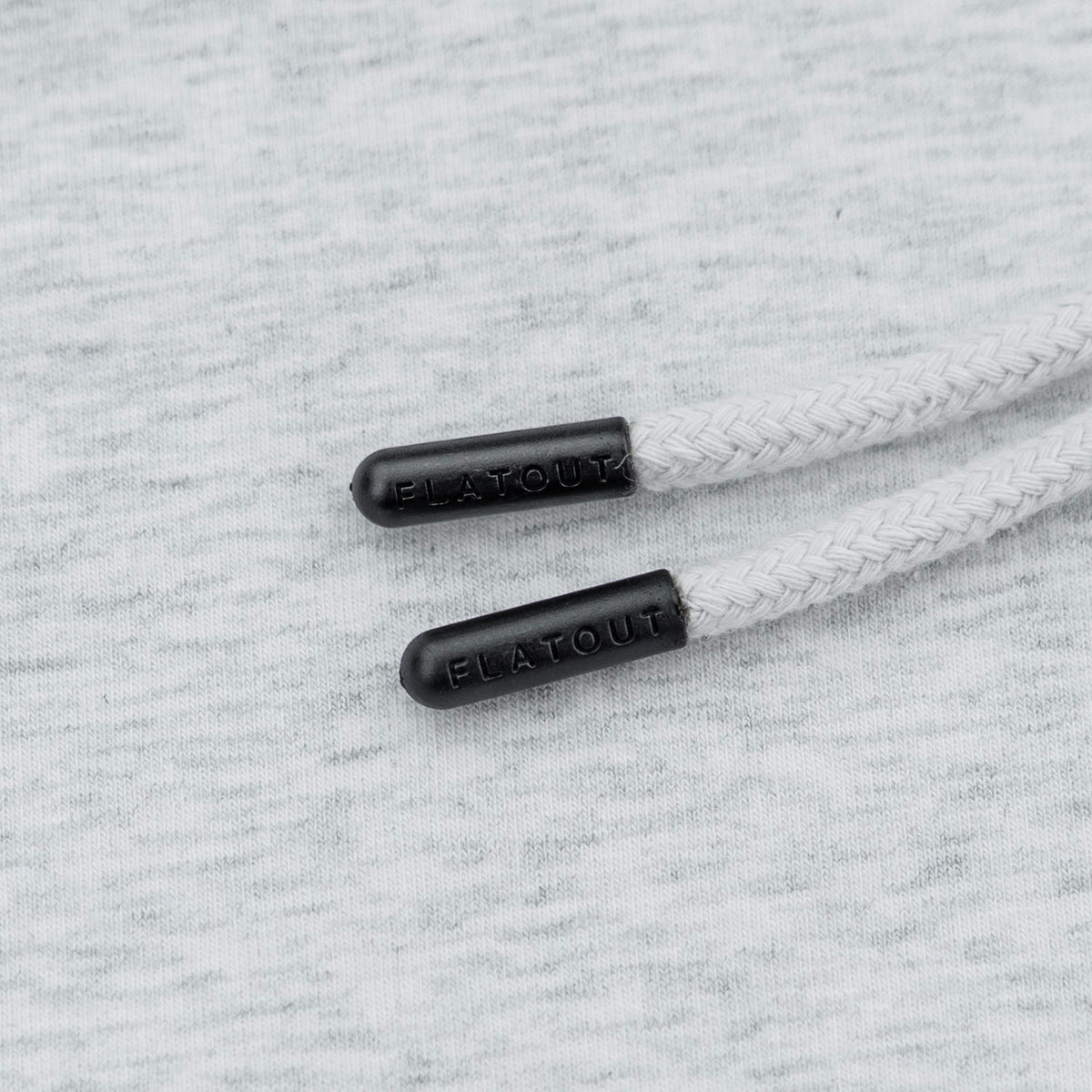Close-up of white braided drawstrings with black plastic tips embossed with the brand name FLATOUT, laid on a light gray heathered fabric background.