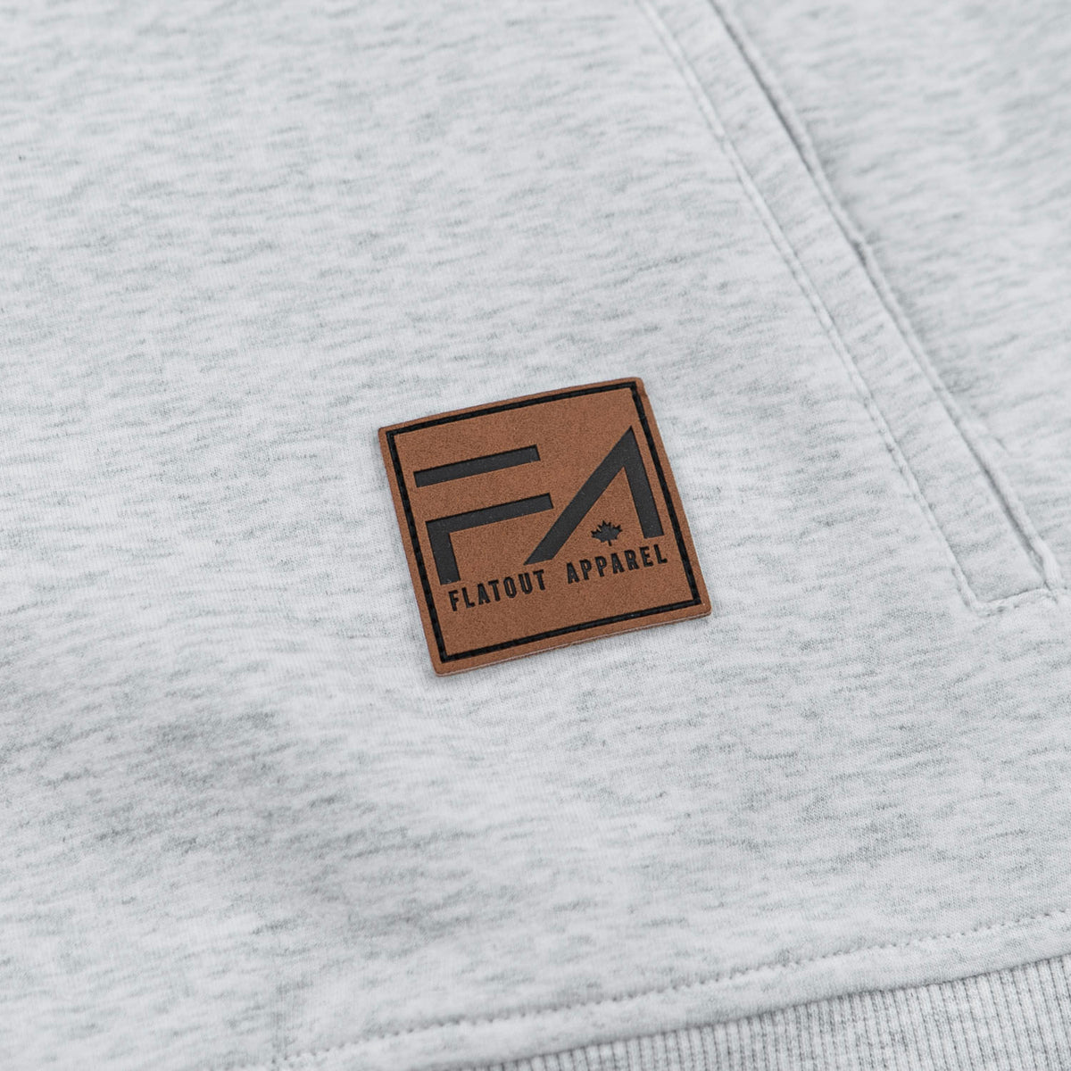 Close-up of a light heather gray sweatshirt featuring a square brown leather patch with the black 'Flaout Apparel' logo on the chest.