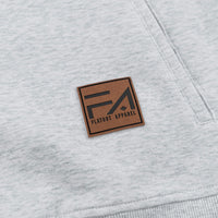 Close-up of a light heather gray sweatshirt featuring a square brown leather patch with the black 'Flaout Apparel' logo on the chest.