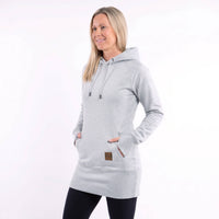 A woman modeling a light heather gray hooded sweatshirt dress with drawstrings, kangaroo pockets, and a small brown brand patch near the lower front, against a plain white studio background.